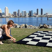 person securing giant chess & checkers mat outdoors next to a lake and city skyline
